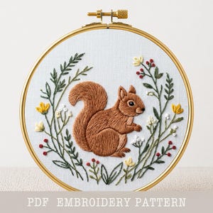 Pode incluir: Um bastidor de bordado detalhado com um esquilo castanho rodeado por folhas verdes, flores amarelas e bagas vermelhas. O desenho está sobre um tecido branco dentro de um aro dourado. O texto "PDF EMBROIDERY PATTERN" está na parte inferior.