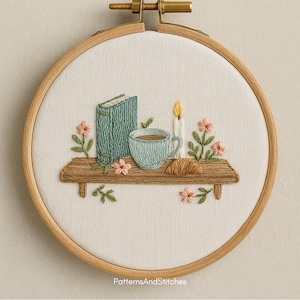 Puede incluir: Arte bordado en un aro de madera. Muestra una estanter&iacute;a de madera con un libro turquesa, una taza de caf&eacute; y un cruas&aacute;n. Tambi&eacute;n est&aacute;n bordadas una vela encendida y flores rosas. Fondo blanco, marco de madera.