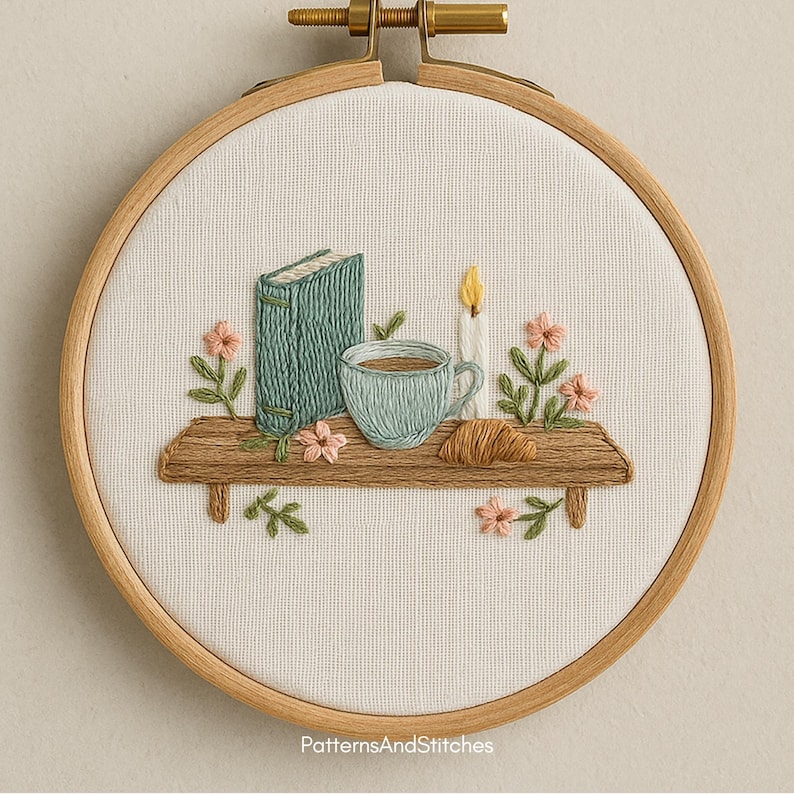 Puede incluir: Arte bordado en un aro de madera. Muestra una estanter&iacute;a de madera con un libro turquesa, una taza de caf&eacute; y un cruas&aacute;n. Tambi&eacute;n est&aacute;n bordadas una vela encendida y flores rosas. Fondo blanco, marco de madera.