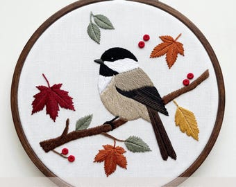 May include: A circular wooden embroidery hoop containing white fabric with a hand-stitched design of a black-capped chickadee perched on a brown branch. The scene includes red, orange, and yellow autumn leaves, green leaves, and small red berries.