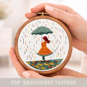 Könnte beinhalten: Ein runder Stickrahmen mit einem gestickten Design einer Person unter einem Regenschirm im Regen. Die Person trägt ein orangefarbenes Kleid und hält einen grünen Regenschirm. Der Text "PDF EMBROIDERY PATTERN" befindet sich unten.