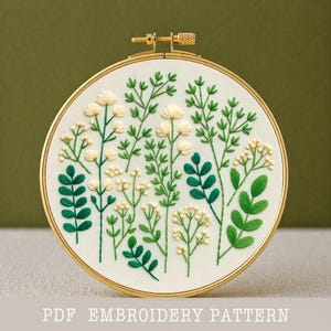 May include: A round embroidery hoop featuring a floral design. The embroidery showcases various green leaves and white flowers on a cream-colored fabric. The hoop is gold-colored, and the text "PDF EMBROIDERY PATTERN" is at the bottom.