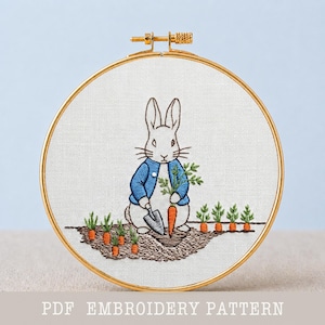 May include: Embroidered hoop art featuring a white rabbit in a blue jacket holding carrots and a shovel in a garden setting. The design includes orange carrots and green plants, all within a gold-colored hoop. Text at the bottom reads "PDF EMBROIDERY PATTERN".