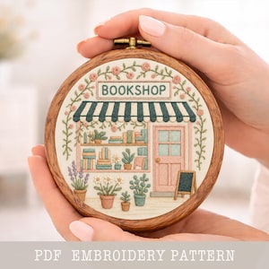 May include: A round, wooden embroidery hoop featuring a detailed embroidered scene of a bookstore. The design includes a green and white striped awning, a pink door, and the word "BOOKSHOP" above. The scene is surrounded by floral and plant details.