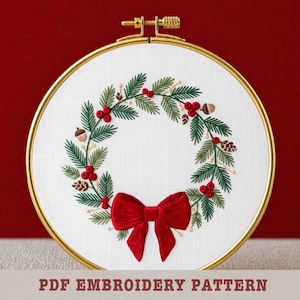 May include: Embroidered wreath in a gold hoop. The wreath features green leaves, red berries, and a red bow. The text "PDF EMBROIDERY PATTERN" is at the bottom. The embroidery is on white fabric.