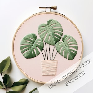 Pode incluir: Padrão de bordado à mão com uma planta em vaso com três folhas verdes de monstera sobre um fundo rosa. O texto "HAND EMBROIDERY PATTERN" está impresso abaixo da planta.