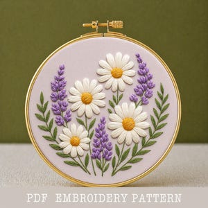 Puede incluir: Un bastidor de bordado floral con margaritas blancas con centros amarillos y ramitas de lavanda morada sobre un fondo lila claro. Hojas verdes y un aro dorado enmarcan el diseño. El texto "PDF EMBROIDERY PATTERN" está en la parte inferior.