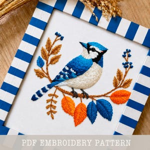 May include: Embroidered artwork of a blue jay on a branch with orange and blue leaves. The bird has detailed blue and white feathers. The artwork is framed in a white and blue striped frame. Text at the bottom reads "PDF EMBROIDERY PATTERN".