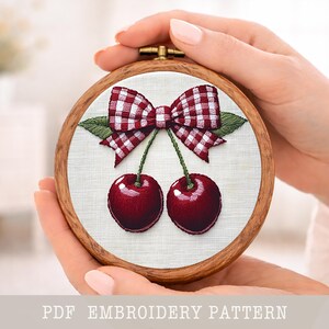Puede incluir: Diseño de cerezas bordadas en un aro de madera. El diseño presenta dos cerezas con un lazo de cuadros rojos y blancos y hojas verdes. El texto "PDF EMBROIDERY PATTERN" está en la parte inferior.