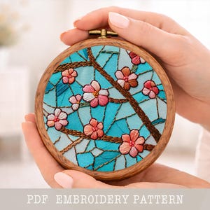 Puede incluir: Un bastidor de bordado de madera redondo con un diseño floral. El diseño incluye flores rosas y blancas, ramas marrones y un fondo de mosaico azul. El texto "PDF EMBROIDERY PATTERN" está en la parte inferior.