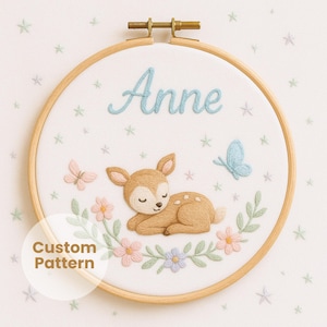 Könnte beinhalten: Gestickte Stickrahmenkunst mit dem Namen "Anne" in hellblauer Stickerei. Ein braunes Reh, Schmetterlinge und Blumen sind unter dem Namen gestickt. Die Worte "Custom Pattern" befinden sich in einem weißen Kreis unten links.