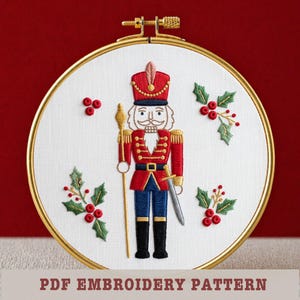 May include: Embroidered hoop art featuring a red and gold nutcracker soldier with a sword and staff, surrounded by holly and berries. The text "PDF Embroidery Pattern" is at the bottom. The hoop is gold-colored, set against a red background.