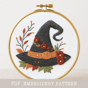 May include: A circular embroidery hoop containing white fabric with a hand-stitched dark gray witch hat design. The hat features an orange band, rust-colored flowers, and green leaves. The text PDF EMBROIDERY PATTERN is printed at the bottom.