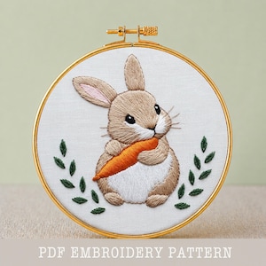 Puede incluir: Un bordado de un conejito marrón y blanco sosteniendo una zanahoria naranja, en un aro dorado. Hojas verdes rodean al conejo. El texto "PDF EMBROIDERY PATTERN" está en la parte inferior.