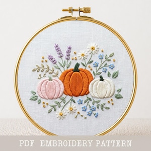 Op de afbeelding: Een ronde borduurring met witte stof en een handgeborduurd ontwerp van drie pompoenen in oranje, roze en wit, omringd door lavendeltakjes, witte madeliefjes en kleine blauwe bloemen. De tekst luidt PDF EMBROIDERY PATTERN.