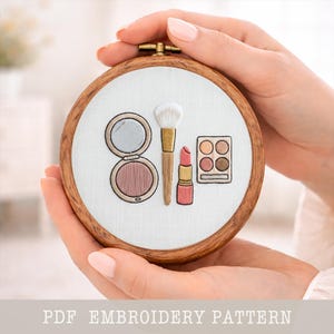 May include: A round embroidery hoop featuring a makeup design. The design includes a compact with a mirror, a makeup brush, a lipstick, and an eyeshadow palette. The text "PDF EMBROIDERY PATTERN" is at the bottom.