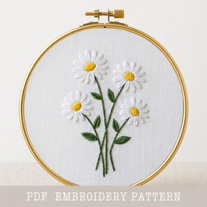 May include: A circular embroidery hoop with a gold-toned frame containing white fabric. The fabric features a hand-stitched design of four white daisies with yellow centers and green stems and leaves. The text PDF EMBROIDERY PATTERN is visible.