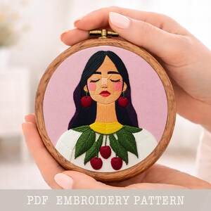 Puede incluir: Un bordado redondo en un aro de madera que representa a una mujer con cabello negro, pendientes de cereza y una camisa blanca con hojas verdes y cerezas rojas. El fondo es rosa claro. El texto "PDF EMBROIDERY PATTERN" está en la parte inferior.