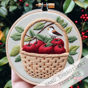 Może przedstawiać: Ręcznie haftowane dzieło sztuki w drewnianym tamborku. Projekt przedstawia kosz wypełniony czerwonymi jabłkami, zielonymi liśćmi i małym ptakiem. Widoczny jest tekst "HAND EMBROIDERY TEMPLATE". Haft jest umieszczony na neutralnej tkaninie.