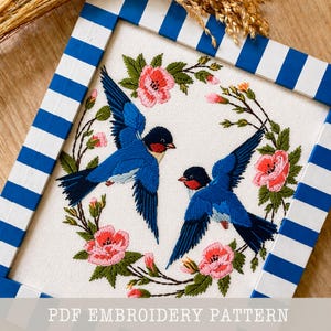 May include: Embroidered artwork featuring two blue birds with red and black accents, surrounded by pink flowers and green leaves. The artwork is framed in a white and blue striped wooden frame. The text "PDF EMBROIDERY PATTERN" is at the bottom.
