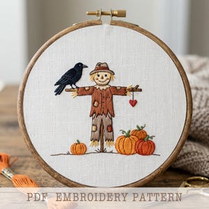 May include: A circular wooden embroidery hoop containing white fabric with a hand-stitched design of a scarecrow wearing a hat, a black crow perched on its arm, a small red heart, and three orange pumpkins at the base. Text reads PDF EMBROIDERY PATTERN.