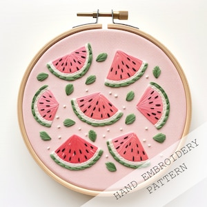 Pode incluir: Padrão de bordado à mão com fatias de melancia e folhas verdes sobre fundo rosa, num bastidor de madeira. O texto "HAND EMBROIDERY PATTERN" é visível. Um objeto artesanal decorativo.