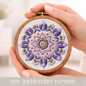 May include: A circular embroidery hoop with a floral pattern. The design showcases a central flower and petals in purple, pink, and green hues. The text "PDF EMBROIDERY PATTERN" is visible at the bottom.