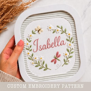 Könnte beinhalten: Weiß gerahmte Stickerei mit dem Namen "Isabella" in rosa Schrift, umgeben von einem Blumenkranz aus weißen Gänseblümchen und grünen Blättern. Ein Schmetterling und eine Libelle sind ebenfalls gestickt. Der Hintergrund hat ein Streifenmuster. Der Text "CUSTOM EMBROIDERY PATTERN" steht unten.