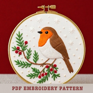 May include: Embroidered robin bird in a gold hoop. The bird has a brown back, orange breast, and white belly, perched on a branch with green leaves and red berries. White snowflakes are scattered around the bird. The text "PDF EMBROIDERY PATTERN" is at the bottom.