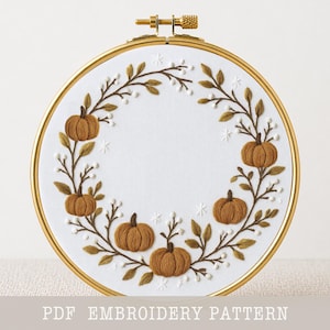 May include: A circular embroidery hoop containing white fabric with a hand-stitched wreath design featuring brown pumpkins, green leaves, and small white berries. The text PDF EMBROIDERY PATTERN is printed at the bottom of the image.