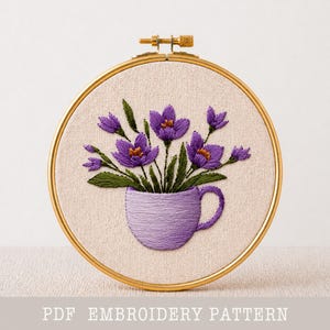 May include: Hand-stitched embroidery art featuring purple flowers in a lavender teacup. The design is set within a gold-colored hoop. The background is a neutral beige fabric. The text "PDF EMBROIDERY PATTERN" is at the bottom.