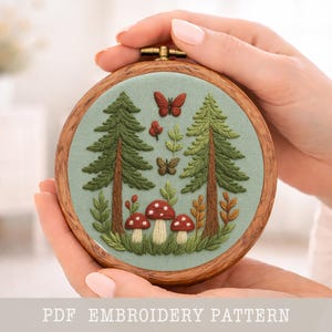 Peut inclure: Un cercle de broderie avec un motif de forêt : deux pins verts, des champignons rouges et des papillons, sur un tissu vert clair. Le cadre est en bois. Le texte en bas indique "PDF EMBROIDERY PATTERN".