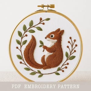 Könnte beinhalten: Gesticktes Eichhörnchen in einem goldenen Reifen. Das Eichhörnchen ist braun mit weißem Bauch und hält ein grünes Blatt. Grüne Blätter und braune Beeren umgeben das Eichhörnchen. Der Text "PDF EMBROIDERY PATTERN" steht unten.