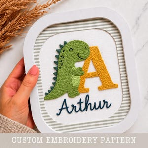 May include: Embroidered artwork featuring a green dinosaur hugging a yellow letter "A". The name "Arthur" is stitched below in navy blue. The design is framed in a white square frame with a striped background. The text "CUSTOM EMBROIDERY PATTERN" is at the bottom.