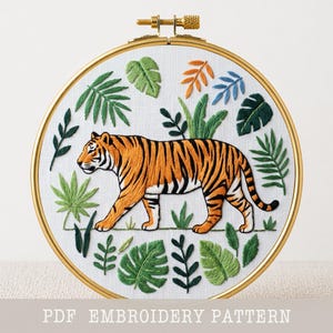 May include: Embroidered tiger in a gold hoop. The tiger is orange and black, surrounded by green and blue leaves. The text "PDF EMBROIDERY PATTERN" is at the bottom of the image.