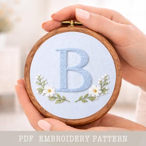 Könnte beinhalten: Ein runder Holz-Stickrahmen mit hellblauem Stoff, auf dem der Buchstabe "B" gestickt ist. Weiße Gänseblümchen und grüne Blätter umgeben den Buchstaben. Der Text "PDF EMBROIDERY PATTERN" befindet sich unten.