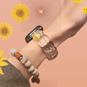 May include: A person's wrist wearing a clear, multi-colored beaded bracelet and a clear, multi-colored watch band. The person is also wearing a brown and white beaded bracelet. The background is a light pink with yellow flowers.