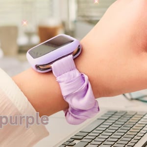Puede incluir: Un reloj inteligente lila con una correa tipo scrunchie a juego. El reloj tiene una pantalla rectangular y una caja redondeada. La correa es de tela fruncida. El texto "Lilac purple" es visible.
