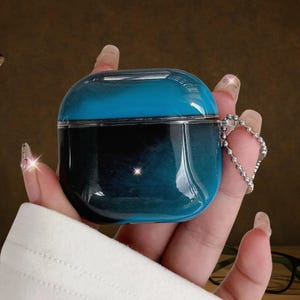 Puede incluir: Una funda protectora azul y negra de dos tonos para auriculares inalámbricos. La funda tiene un acabado brillante y transparente y una cadena plateada para sujetarla. La funda se sostiene en una mano con uñas largas y cuidadas.