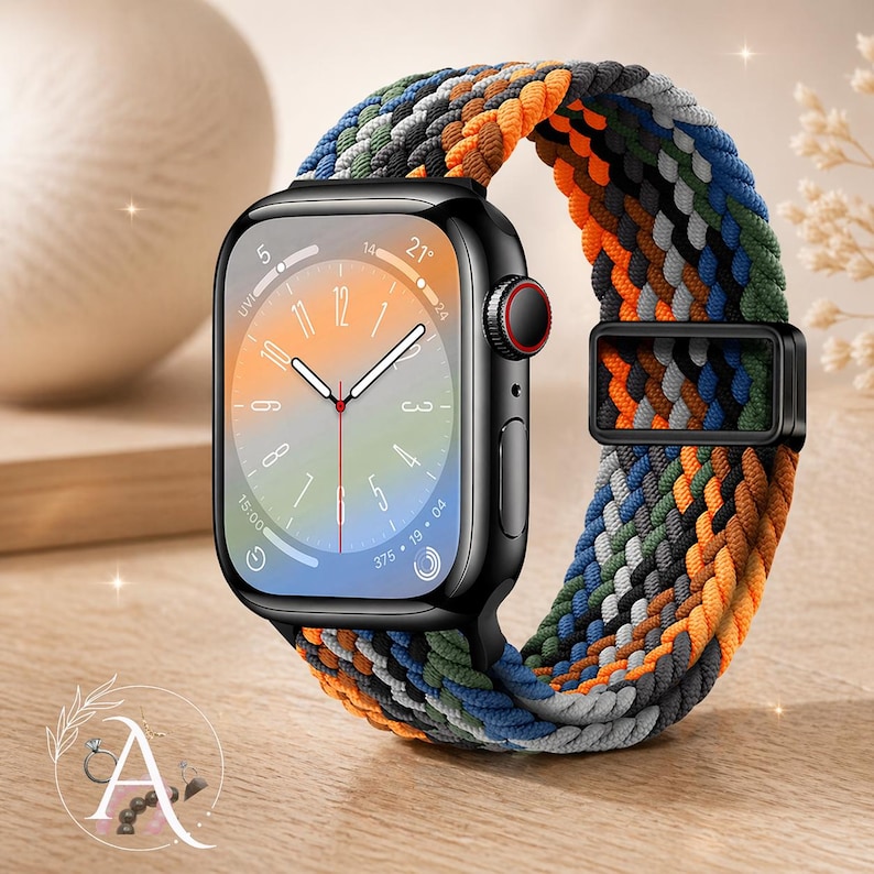 May include: A black smartwatch with a colorful braided band in shades of orange, blue, green, and gray. The watch face displays the time. The band is secured with a black metal clasp. The watch is on a wooden surface.