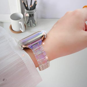 Puede incluir: Una correa y una caja de reloj transparentes con un acabado iridiscente y brillante. La correa está hecha de eslabones rectangulares, y la caja tiene un marco metálico. El reloj se lleva en la muñeca, mostrando su diseño colorido y llamativo.