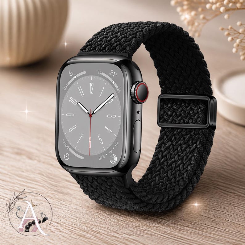 May include: A black smartwatch with a braided black band. The watch face displays the time and other information. The band is secured with a black metal clasp. The watch is on a wooden surface.