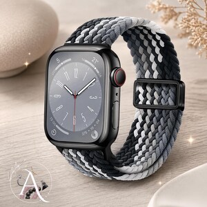 May include: A black smartwatch with a gray watch face and a braided watch band in shades of black, gray, and white. The watch band is secured with a black metal clasp. The watch is displayed on a light-colored wooden surface.