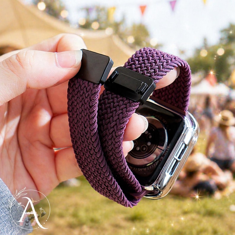 May include: A deep purple braided Apple Watch band with a black clasp. The band is held up, showing the woven texture and the watch face. The watch has a clear case.
