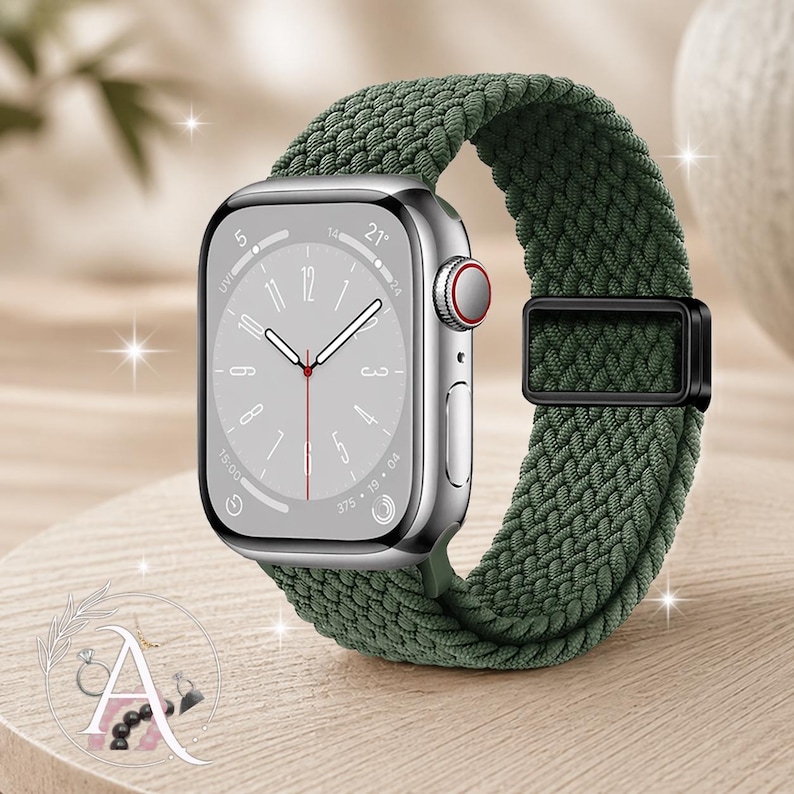 May include: A silver smartwatch with a green braided band. The watch face displays the time and other metrics. The band is secured with a black metal clasp. The watch is displayed on a light brown surface.