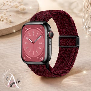 May include: A black smartwatch with a burgundy braided band. The watch face is burgundy with white hands and numerals. The band has a black metal buckle. The watch is displayed on a light wood surface.