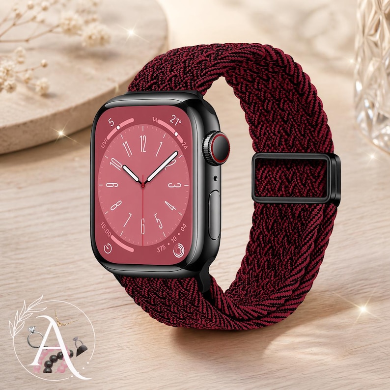 May include: A black smartwatch with a burgundy braided band. The watch face is burgundy with white hands and numerals. The band has a black metal buckle. The watch is displayed on a light wood surface.