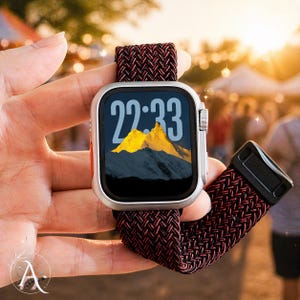 May include: A silver smartwatch with a black screen displaying the time 22:33 and a mountain landscape. The watch has a red button and a burgundy and black woven band. The watch is held in a hand.