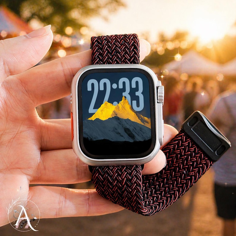 May include: A silver smartwatch with a black screen displaying the time 22:33 and a mountain landscape. The watch has a red button and a burgundy and black woven band. The watch is held in a hand.