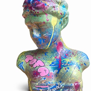 Puede incluir: Busto pintado de una figura clásica, con una base verde y vibrantes salpicaduras de pintura rosa, azul y amarilla. El busto presenta letras rosas de estilo grafiti en el pecho y la firma del artista en la base.